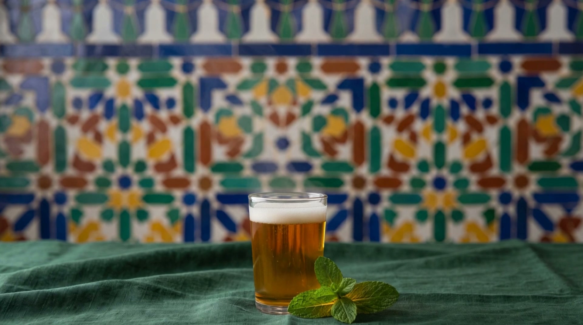 A full glass of mint tea with a sprig of mint on a green tablecloth, in front of a colorful zellige wall.